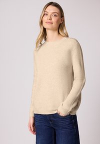 Femme portant un pull en maille beige avec des boutons sur l'épaule et un jean bleu foncé, debout, une main dans la poche, sur un fond uni.