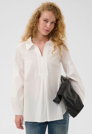 Vrouw met krullend blond haar die een losse witte blouse en blauwe spijkerbroek draagt en een zwart leren jack vasthoudt tegen een effen achtergrond.