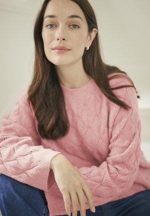 Pulover tricotat roz cu un model texturat în diamant, purtat cu blugi casual albaștri. Atmosfera este suavă și neutră.