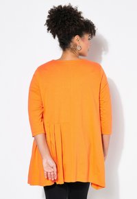 Orangene Tunika mit 3/4-Ärmeln, plissiertem Rückendesign, rundem Ausschnitt, weichem Stoff und einem etwas längeren Saum hinten.
