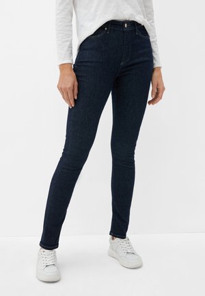 Persoon draagt donkerblauwe skinny jeans, een wit lang shirt met lange mouwen en witte sneakers, staand tegen een effen lichte achtergrond.