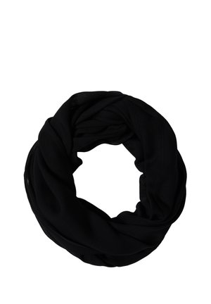 Lachende vrouw die een wit T-shirt met korte mouwen, een zwarte sjaal en blauwe jeans draagt, met één hand in de zak tegen een neutrale achtergrond.