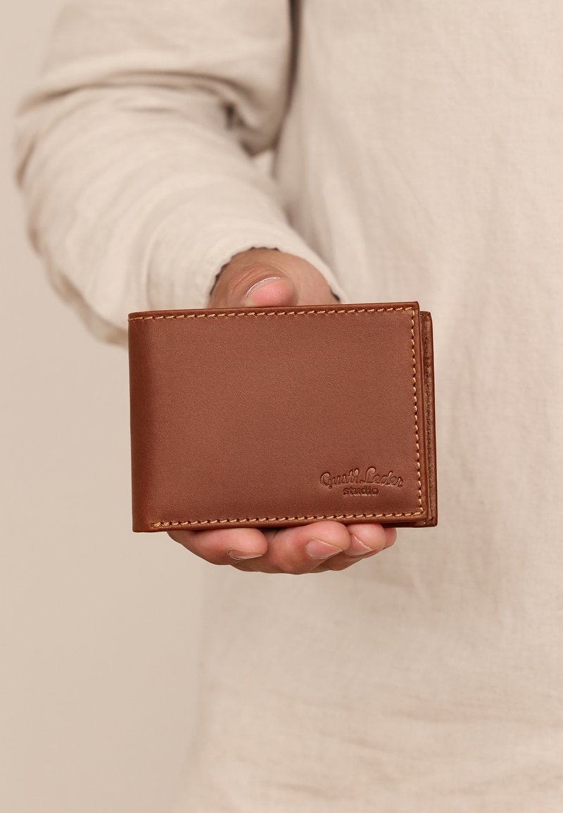 Brązowy skórzany portfel o gładkiej teksturze, w układzie bi-fold, z widocznym przeszyciem i wytłoczonym logo na powierzchni. Kompaktowy rozmiar do trzymania w ręce.