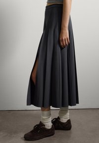Donna che indossa una lunga gonna plissettata scura con uno spacco laterale, calze bianche, sneakers scure e un top grigio corto davanti a un muro neutro.