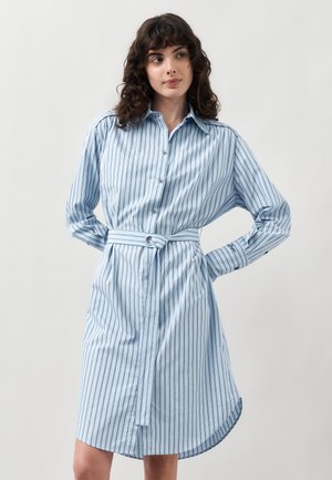 Kobieta ubrana w jasnoniebieską, długą sukienkę z guzikami, w pionowe paski i z paskiem w talii, stoi z rękami za plecami.