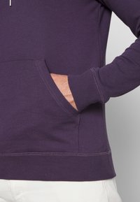 Felpa viola con tasca a marsupio, polsini e orlo a costine. Il materiale sembra morbido e con una texture liscia. Design semplice e funzionale.
