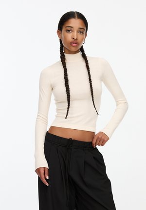 Pulover turtleneck de culoare bej deschis, dintr-un material tricotat în relief, cu o siluetă ajustată, mâneci lungi, cu orificii pentru degete. Asortat cu pantaloni negri largi.