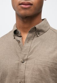 Svetlohnedá košeľa na gombíky s ostrým golierom, s náprsným vreckom a textúrovanou tkaninou. Současťou sú kontrastné gombíky na lége.