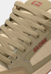 Gros plan sur les lacets beiges d'une basket, le panneau latéral avec le logo rouge "GLOBE", et les coutures détaillées sur les matériaux en tissu et cuir.