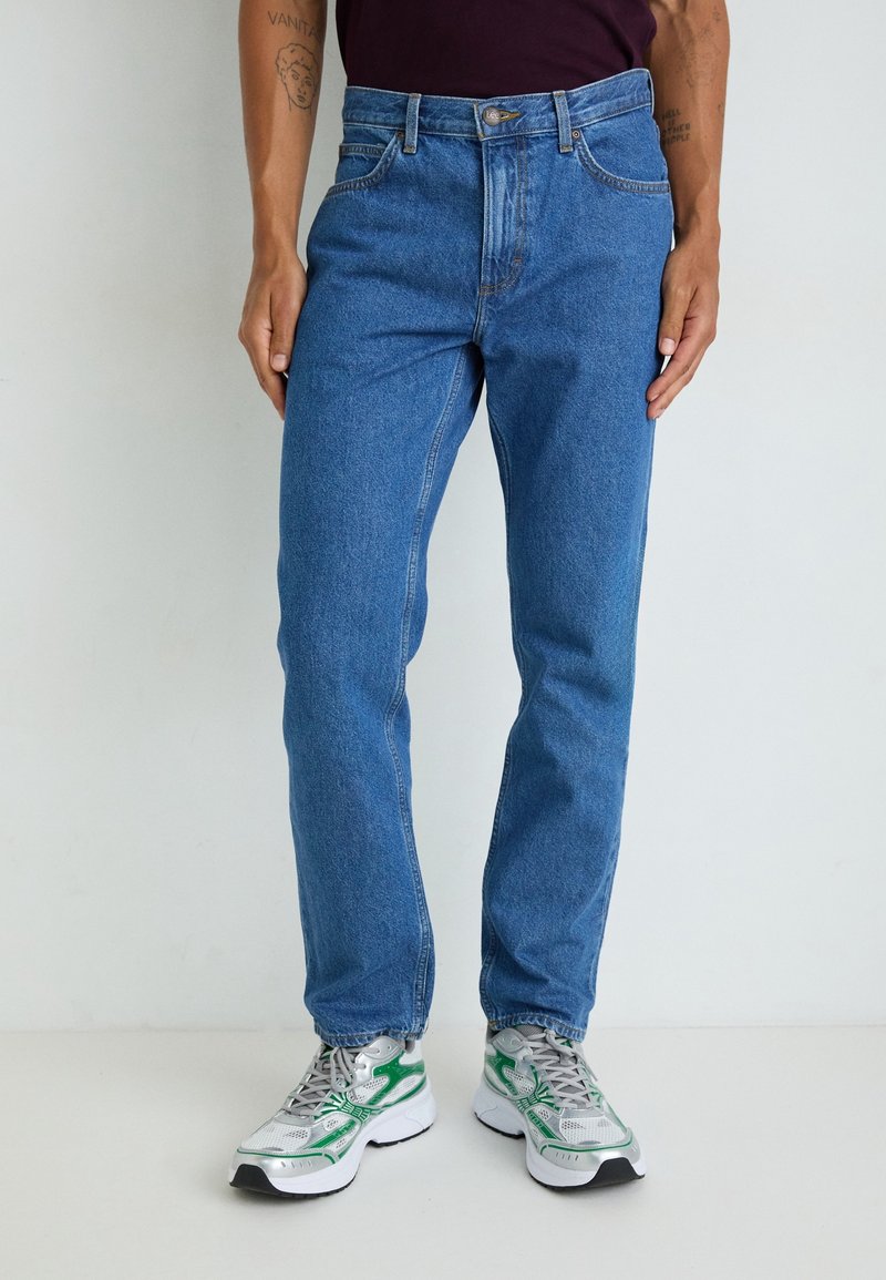 Personne portant un jean bleu taille moyenne à jambes droites avec poches avant et des baskets blanches avec des accents verts, debout devant un fond uni.