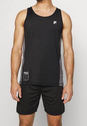 Mężczyzna ubrany w czarny sportowy tank top bez rękawów i pasujące czarne szorty stojący na jednolitym tle.