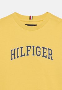 Κίτρινο βαμβακερό μπλουζάκι με στρογγυλή λαιμόκοψη και λογότυπο "HILFIGER" σε μπλε ναυτικό χρώμα τυπωμένο στο στήθος. Διαθέτει ριγωτή λεπτομέρεια στη λαιμόκοψη.