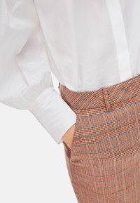 Weißes Hemd mit Puffärmeln, kombiniert mit braunen karotypierten Hosen mit blauen Akzenten. Die Hose hat ein Detail an der Vordertasche.