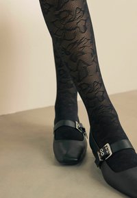 Zwarte panty's met een bloemmotief in kant, gecombineerd met zwarte platte schoenen met een vierkante neus en een metalen gesp als accent.