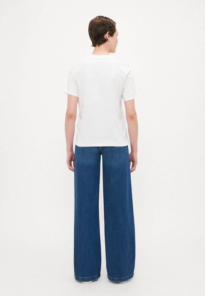 Weißes T-Shirt mit kurzen Ärmeln, kombiniert mit weiten blauen Jeans, die einen lockeren Schnitt und ein minimalistisches Design aufweisen, von hinten betrachtet.