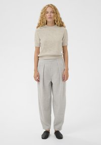 Femeie cu păr lung, ondulat, blond, îmbrăcată cu un top tricotat bej cu mânecă scurtă, pantaloni gri deschis plié și pantofi negri tip slip-on, stând pe un fundal alb.