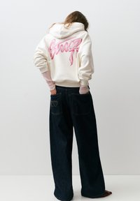 Persoană purtând un hanorac crem cu text roz în stil graffiti pe spate, suprapus peste mâneci roz, asortat cu blugi evazați închis la culoare cu buzunar brodat.