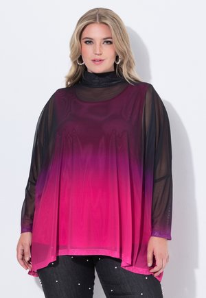 Blonde vrouw die een doorschijnend zwarte top met lange mouwen draagt onder een losse, roze tot paarse verloop mouwloze tuniek en een zwarte broek met parels.