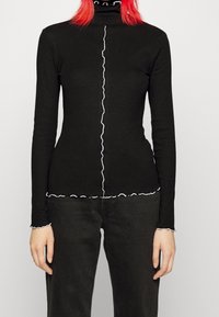 Svart långärmad polotröja med vita volanger längs kragen, knäppslån och nederkant. Slim fit-design, stickad tyg.