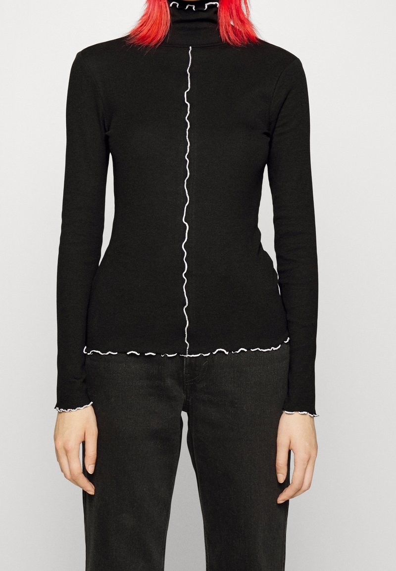 Svart långärmad polotröja med vita volanger längs kragen, knäppslån och nederkant. Slim fit-design, stickad tyg.