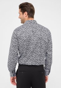 Uomo di spalle che indossa una camicia a maniche lunghe bianca e nera con un motivo e pantaloni neri su uno sfondo bianco uniforme.