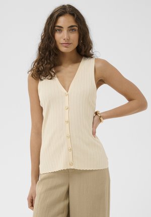 Kvinde med krøllet hår iført en ærmeløs cremefarvet knapbluse og beige bukser, stående med den ene hånd på hoften mod en ensfarvet baggrund.