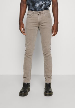 Άνδρας που φοράει στενά μπεζ τζην, μαύρες δερμάτινες μπότες και ένα σκοτεινό πουκάμισο tie-dye، στέκεται έναντι μιας απλής ανοιχτόχρωμης φόντου.