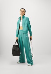 Τιρκουάζ φόρμα με λευκές ρίγες, περιλαμβάνει τζάκετ με φερμουάρ και παντελόνι με ελαστική μέση. Συμπεριλαμβάνονται μαύρη αθλητική τσάντα και ρακέτα τένις.