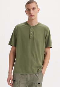 Olivovo zelené tričko s krátkym rukávom typu henley s päťgombíkovou légiou. Mäkká bavlnená látka s voľným strihom a okrúhlym výstrihom.