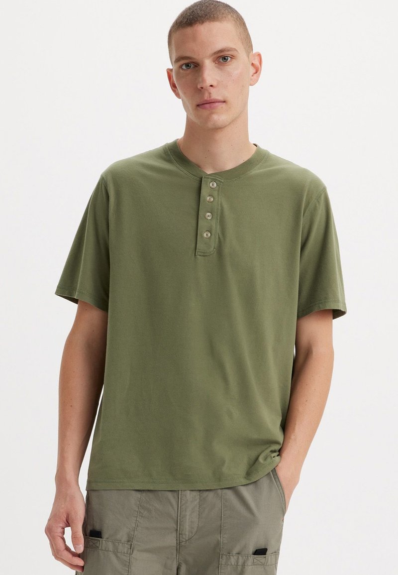 Olivovo zelené tričko s krátkym rukávom typu henley s päťgombíkovou légiou. Mäkká bavlnená látka s voľným strihom a okrúhlym výstrihom.