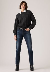 Γυναίκα με σκούρο γκρι πουλόβερ με πλέξιμο καλωδίου πάνω από λευκό πουκάμισο με γιακά, μπλε τζιν, μαύρη ζώνη και μαύρες μπότες στον αστράγαλο, που στέκεται μπροστά σε απλό φόντο.