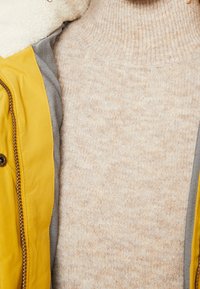 Beige stickad turtlenecktröja under en gul jacka med synlig dragkedja och vitt fleecefoder.