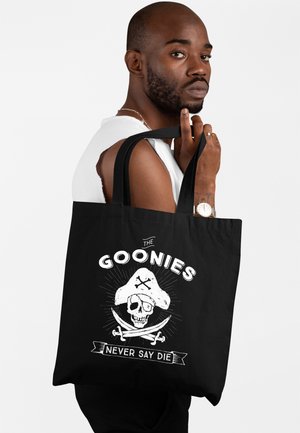 Mężczyzna z brodą w białym bezrękawnym t-shircie trzyma czarną torbę z białym pirackim czaszką i napisem "The Goonies Never Say Die".