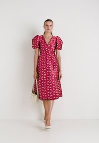 Vestido envolvente fúcsia com mangas bufantes, padrão floral, até ao joelho, feito de tecido suave. Modelo segurando uma pequena bolsa branca.