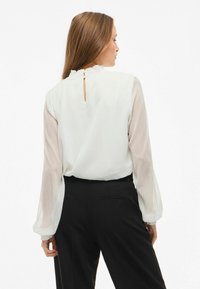 Hvid bluse med gennemsigtige lange ærmer, rund hals og plisseret detaljer bagpå, parret med sorte højtaljede bukser.