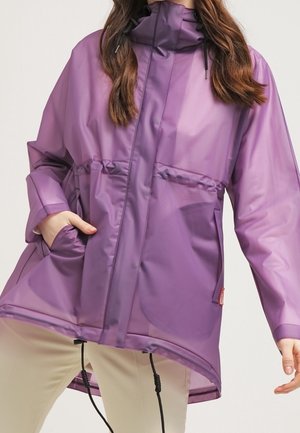 Kobieta ubrana w luźną, fioletową kurtkę przeciwdeszczową z troczkami, przednimi kieszeniami oraz beżowymi spodniami na jednolitym tle.