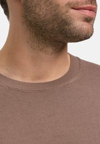 Brauner Strickpullover mit geripptem Rundhals-Ausschnitt, glatter Textur und subtiler Schimmerung. Nahaufnahme des Halsbereichs und kurzen Gesichtshaars.