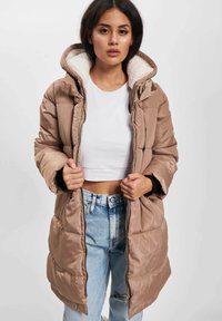 Kvinne som har på seg en beige puffjakke med hvit fleecehjelmfôr, hvit crop-top og lyseblå jeans, mens hun holder jakken åpen ved glidelåsen.