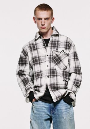 Jeune homme aux cheveux courts portant une chemise à carreaux noire et blanche par-dessus un haut noir et un jean bleu déchiré, les mains dans les poches, fond neutre.