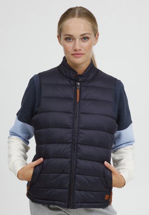 Gilet matelassé bleu marine avec un col haut, une fermeture éclair à l'avant et des poches latérales. Présente des manches en tissu contrasté et un accent en cuir près de l'ourlet.