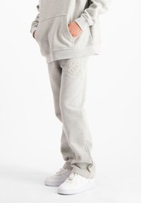 Persoon in lichtgrijze sweatpants en bijpassende hoodie, met handen in de zakken, gecombineerd met witte sneakers, staande tegen een witte achtergrond.