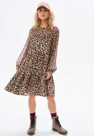 Robe à imprimé léopard avec des manches longues et transparentes et une jupe à volants. Assortie à des bottes de combat imprimé léopard. Palette de couleurs : tons beige et marron.