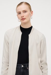 Béžová bomber bunda s černým žebrovaným rolákem pod ní. Bunda má zip, volný střih a hladkou texturu. Tmavé džínové kalhoty.