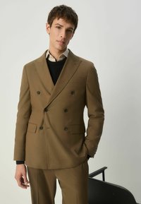 Brūns divrindu uzvalks ar izvirzītām atloks, divām priekšējām kabatām un melniem pogām. Nēsāts pār melnu džemperi un baltu kreklu. Pielāgota piegriezums.