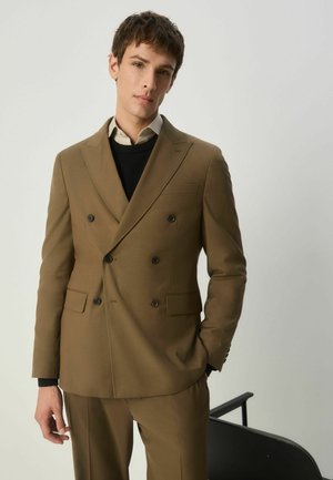 Brauner zweireihiger Anzug mit Spitzrevers, zwei Fronttaschen und schwarzen Knöpfen. Getragen über einem schwarzen Pullover und einem weißen Hemd. Figurbetonte Passform.