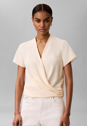 Kobieta z ciemnymi włosami związanymi w niski kok nosi kremową bluzkę kopertową z krótkim rękawem i białe spodnie na jednolitym szarym tle.