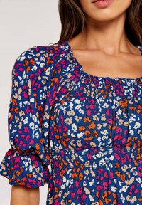 Bluse mit Blumenmuster, die einen marineblauen Hintergrund mit rosa, orange und weißen Blumen zeigt. Rüschenausschnitt und kurze Puffärmel. Weicher Stoff.