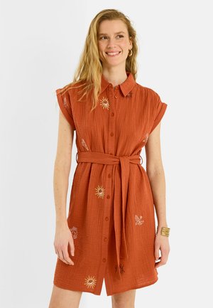 Mujer sonriente con vestido de manga corta color óxido con botones al frente, cinturón en la cintura y bordado de sol y pájaro, complementado con pendientes de aro dorados y pulsera.
