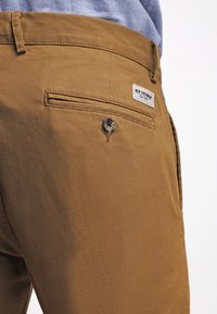 Beige Baumwollhose mit glatter Textur. Verfügt über eine geknöpfte Gesäßtasche und ein darüber angenähtes Etikett mit dem Markennamen.