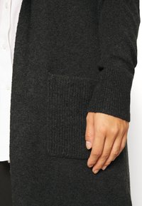 Černý pletený kardigan s texturovaným povrchem, žebrovanými manžetami a kapsou vpředu na levé spodní straně.